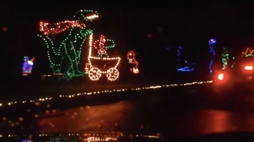 Colorful holiday lights display featuring a dinosaur, a cart, and various festive characters along a dark road.