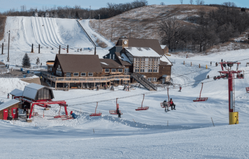 A snowy ski resort with a lodge, ski lifts, and people skiing on the slopes under a clear blue sky.