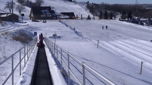 A snowy hill with a conveyor belt for sledding, people walking in the distance, and a lodge in the background.