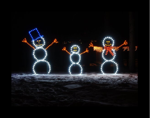 Three illuminated snowmen with a blue hat, orange arms, and a red scarf, standing in a snowy landscape at night.