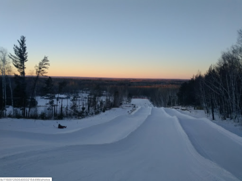 A snowy landscape at dusk, with gentle slopes and trees silhouetted against a colorful horizon.