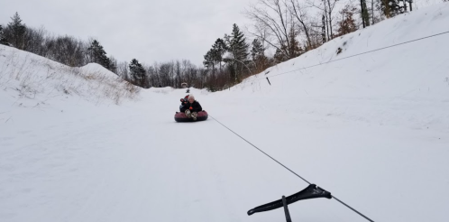 A person tubing down a snowy slope, being pulled by a rope, surrounded by trees and winter scenery.