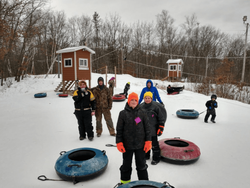 A group of children and adults in winter gear stand on a snowy hill with inflatable tubes and a small building in the background.