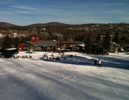 A snowy ski resort with people skiing and snowboarding, surrounded by trees and mountains under a clear blue sky.