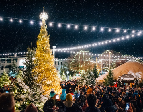 A festive crowd gathers around a large, illuminated Christmas tree amidst falling snow and twinkling lights.
