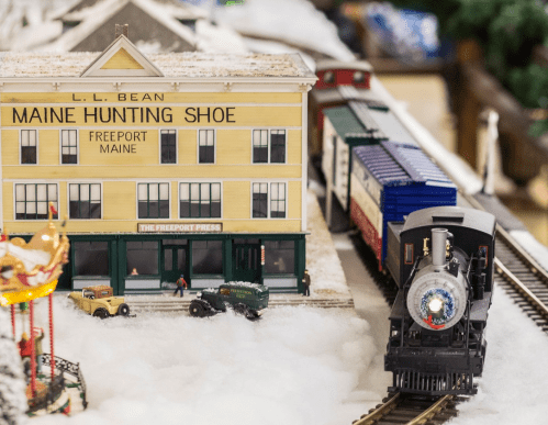 A miniature train set with a model of L.L. Bean's Maine Hunting Shoe store and vintage cars on a snowy landscape.