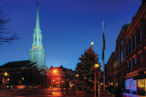 A festive scene featuring a church steeple, a decorated Christmas tree, and buildings illuminated at dusk.