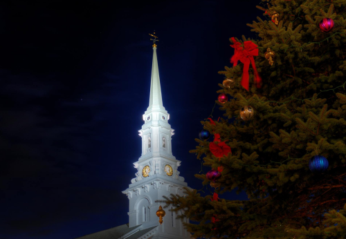 A lit church steeple at night beside a decorated Christmas tree with ornaments and ribbons.