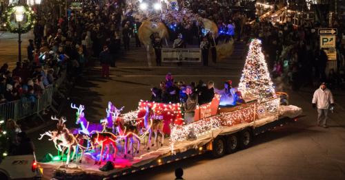 A festive parade scene with a decorated float featuring reindeer and a Christmas tree, surrounded by a cheering crowd.