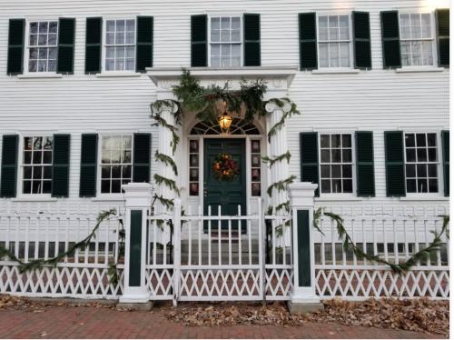 A festive white house with green shutters, decorated with garlands and a wreath on the door, surrounded by a white fence.