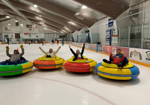 Four people in colorful inflatable tubes wave happily on an ice rink, surrounded by a sports facility.