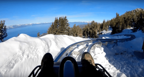 A view from a sled on a snowy track, overlooking a lake and mountains under a clear blue sky.