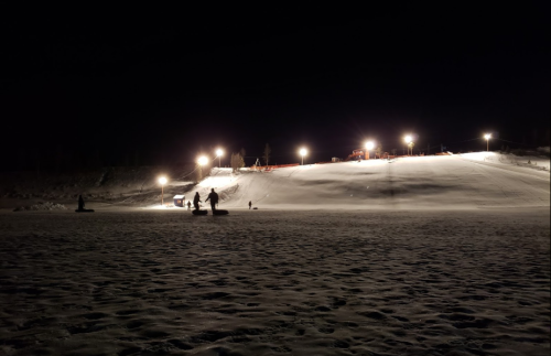 A snowy hill at night, illuminated by bright lights, with people enjoying winter activities in the foreground.