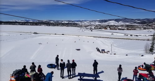 A snowy landscape with people tubing down a hill, surrounded by mountains and a clear blue sky.