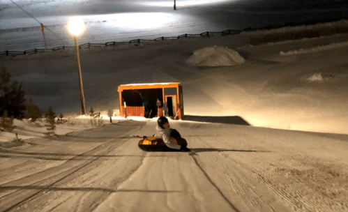 A person sledding down a snowy hill at night, with a lit cabin in the background and a clear sky.