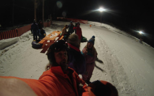 A group of people in winter gear at a snow tubing area at night, with snow-covered ground and lights in the background.