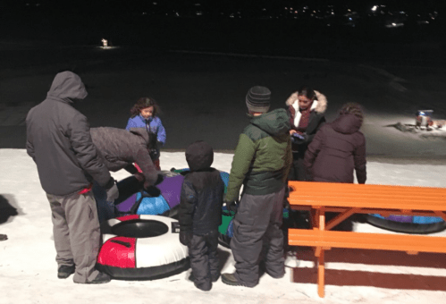 A group of children and adults in winter clothing gather around colorful snow tubes on a snowy hill at night.