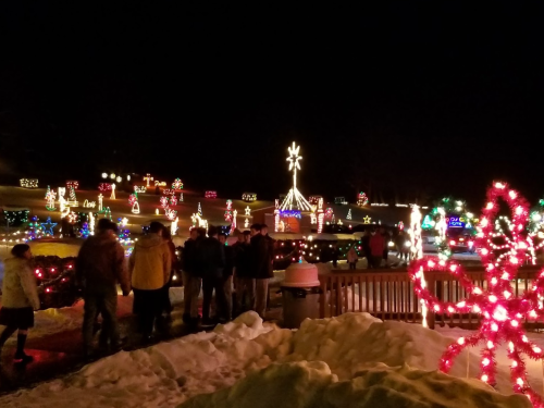 A festive scene at night with colorful holiday lights, decorations, and people walking through a snowy area.