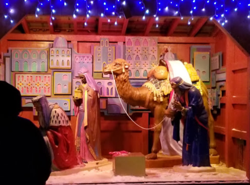 Nativity scene featuring figures of the Magi, a camel, and a backdrop of colorful buildings, illuminated by lights.