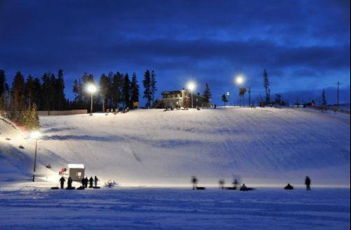 A snowy hill at dusk, with skiers and snowboarders illuminated by lights, surrounded by trees.