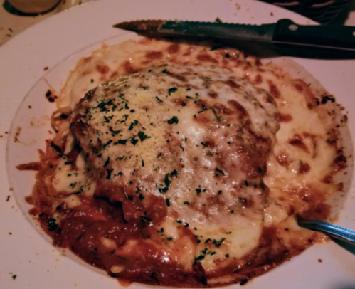 A plate of cheesy baked pasta with a rich tomato sauce, garnished with herbs. A knife and fork are nearby.