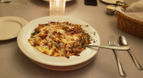 A plate of cheesy baked pasta topped with herbs and bacon, served on a table with utensils and a candle in the background.