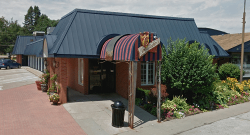 A brick building with a blue metal roof and striped awning, surrounded by colorful flowers and greenery.