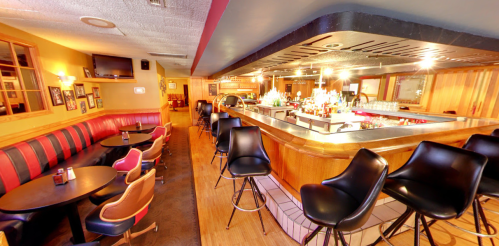 A cozy bar interior featuring a long counter, high stools, and red leather seating, with warm lighting and wooden accents.