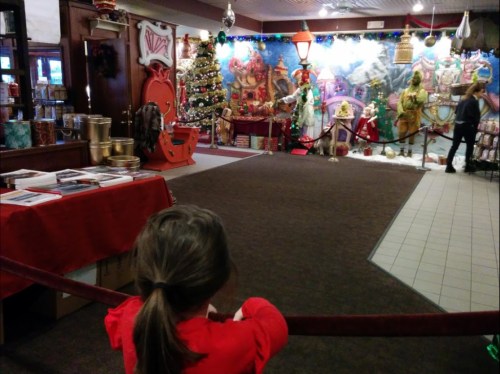 A child in a red sweater looks at a festive holiday display with decorations, a Christmas tree, and costumed figures.