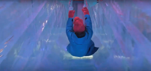 A person in a blue coat and red gloves slides down an icy tunnel illuminated with colorful lights.
