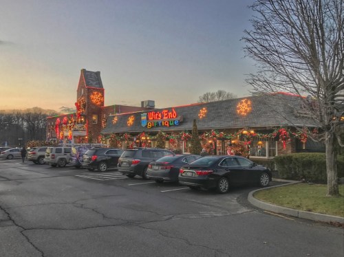 A festive storefront decorated with holiday lights and ornaments, surrounded by parked cars at dusk.