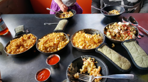 A table with several small bowls of macaroni and cheese, accompanied by sauces and a child's hand reaching for food.