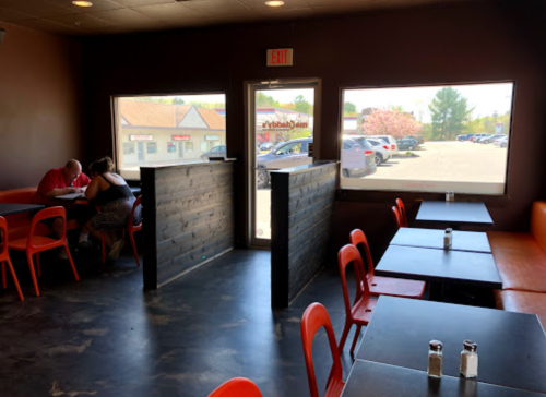 Interior of a casual restaurant with orange chairs, black tables, and large windows showing a parking lot outside.