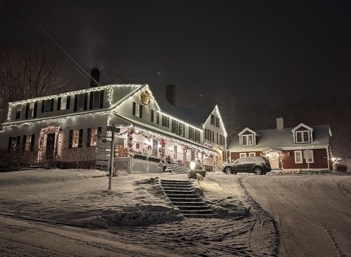 A snow-covered house decorated with lights at night, with a car parked nearby and a cozy atmosphere.