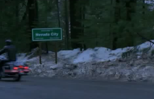 A motorcycle passes a sign for Nevada City, surrounded by trees and patches of snow on the roadside.