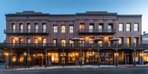 Historic brick building with balconies and large windows, illuminated at dusk, featuring outdoor seating.