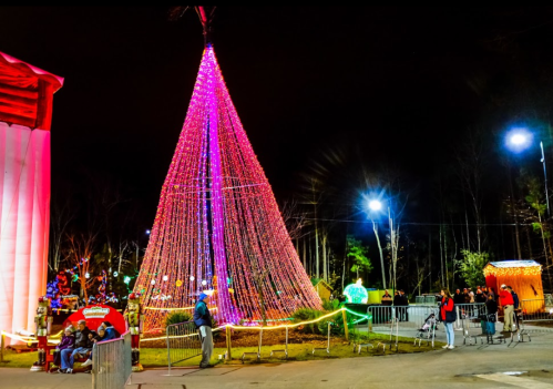 A brightly lit Christmas tree made of lights stands in a festive park, with people enjoying the holiday atmosphere.