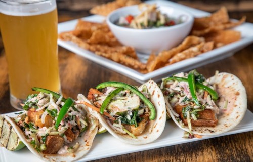 Three tacos filled with meat, greens, and peppers, served with a side of chips and a bowl of salsa, alongside a beer.