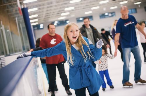 A girl in a blue coat smiles while ice skating, with people skating and holding hands in the background.