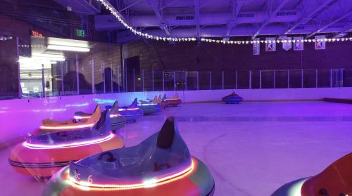 Colorful bumper cars on an ice rink, illuminated by vibrant lights, with a spacious arena in the background.