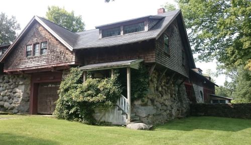 A rustic house with a stone foundation, wooden shingles, and lush greenery surrounding the entrance.