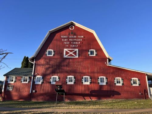 Red barn with white trim, featuring the text "Three Cedars Farm" and names of the owners, set against a clear blue sky.