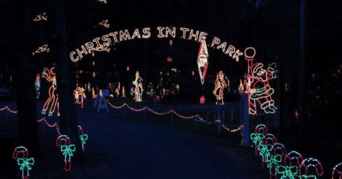 A festive display of Christmas lights in a park, featuring a sign that reads "Christmas in the Park" and holiday decorations.