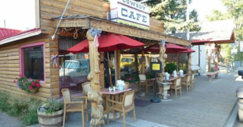 A rustic wooden café with red umbrellas and outdoor seating, surrounded by flowers and a welcoming sign.