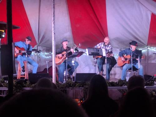 Four musicians play acoustic guitars on stage under a red and white tent, with lights in the background.