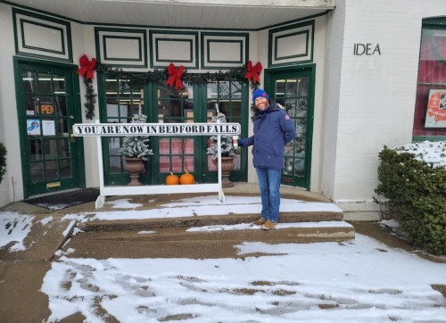 A person stands next to a sign reading "You are now in Bedford Falls" outside a decorated storefront in winter.