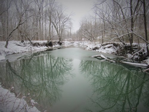 A tranquil river reflects bare trees and snow-covered banks in a serene winter landscape.
