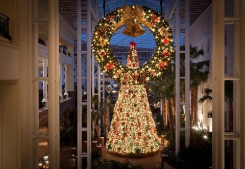 A beautifully decorated Christmas tree surrounded by a large festive wreath and twinkling lights in a spacious atrium.
