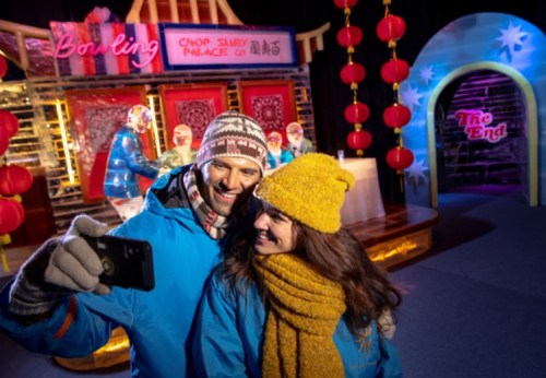 A couple in winter attire smiles while taking a selfie in a festive, colorful setting with decorations and lights.