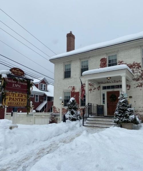 A snowy scene of the Butler House Inn, featuring a brick exterior, festive decorations, and a welcoming entrance.